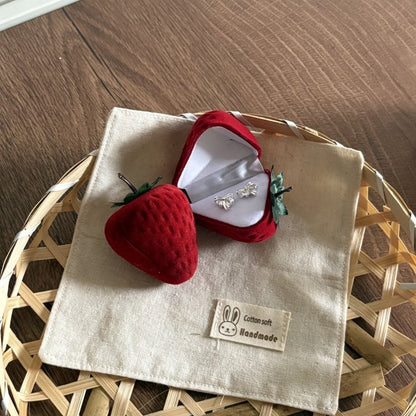 A strawberry-shaped red velvet ring box opened to reveal silver stud earrings, placed on a beige fabric pouch with a bunny patch, set on a woven tray over a wood surface.