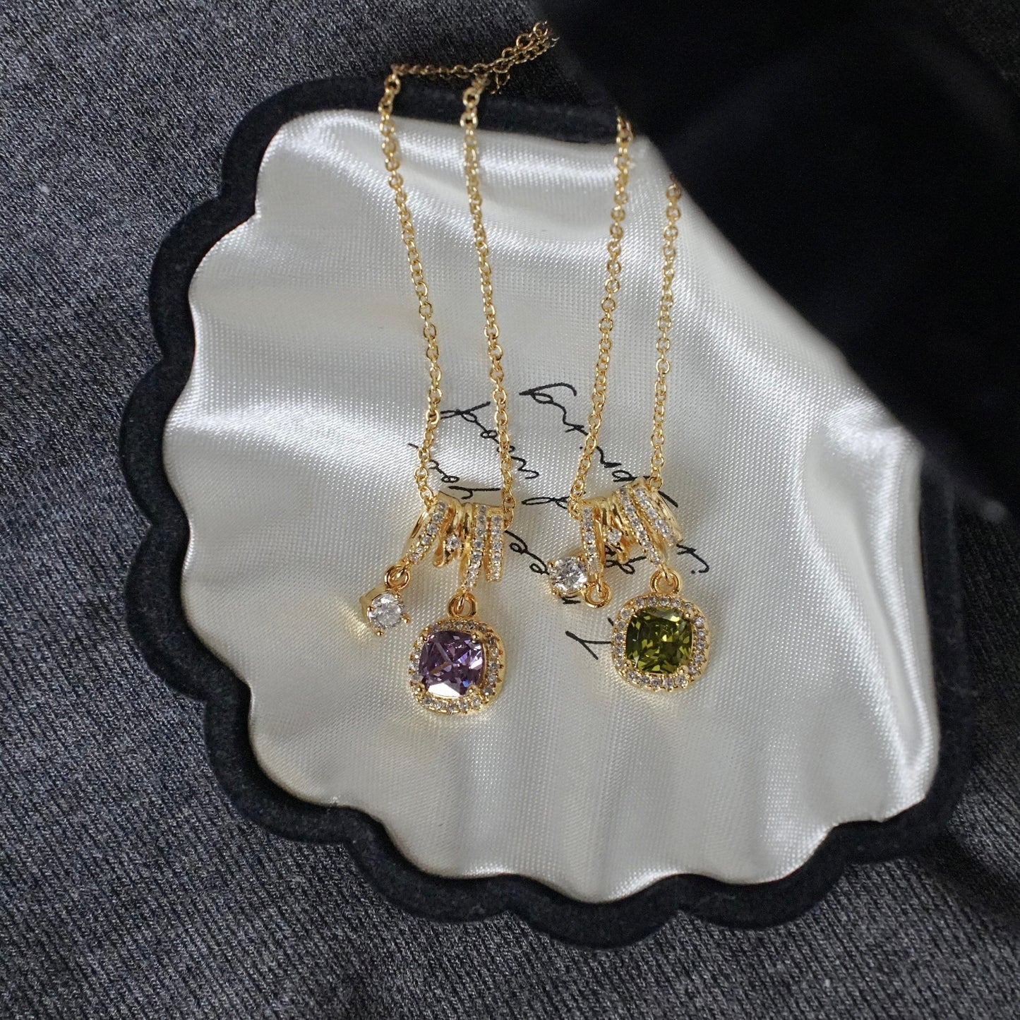 Two gold pendant necklaces displayed on a scalloped white satin card with a black border, set against a gray textured fabric background. One pendant features a purple gemstone and the other features an olive green gemstone, both in gold settings.